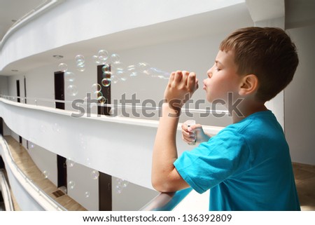 Boy in blue t-shirt lets bubbles on balcony inside large hotel.