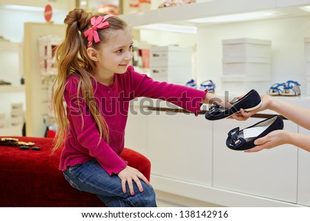 Little girl sitting on ottoman, takes pair of shoes from hands of seller