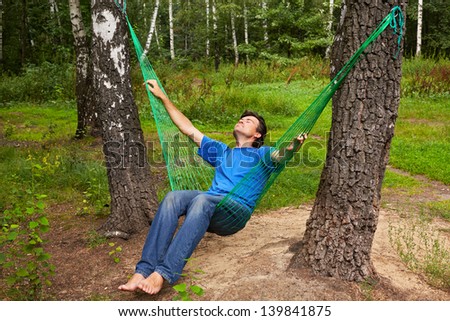 Barefooted man sits in hammock suspended between two thik birches, head thrown back, eyes closed