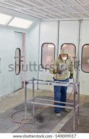 Man in protective clothes and respirator works in paint-spraying booth, painting car details with airbrush