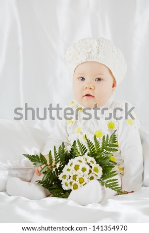 Portrait of little girl in white clothes, who sits on white coverlet between pillows with bunch of flowers
