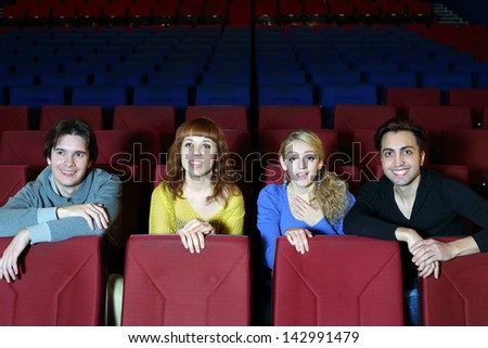 Four young smiling friends sit on seats in cinema theater and see movie.