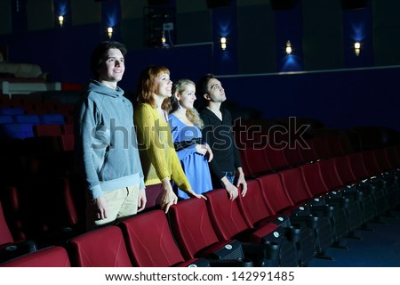 Four happy friends stand and look at screen in cinema theater. Focus on left pair.