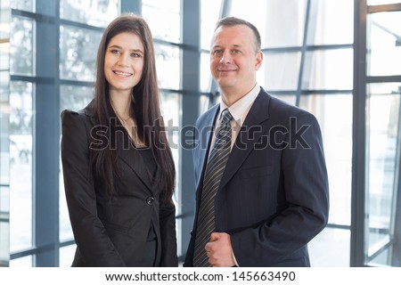 Smiling man and woman in business suits stand near the glass windows