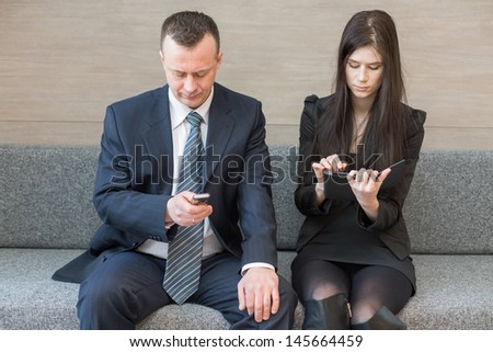 Man and woman in business suits sitting on the couch with a communication device, focus on a man.