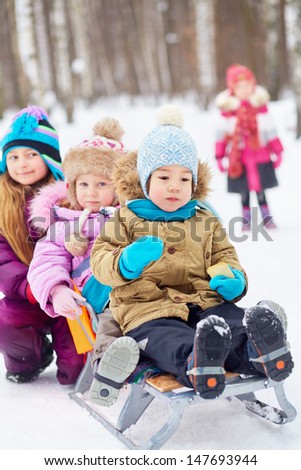 Older girl pushes sled with two little children, first asian boy eats cracker, another one girl stands behind not in focus