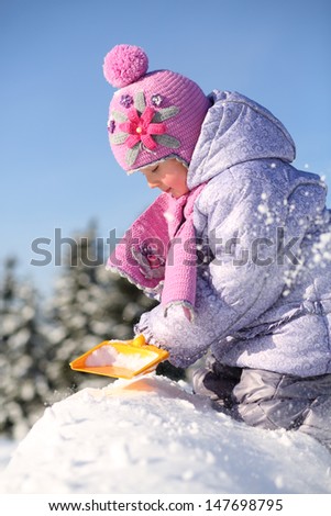 Little girl dressed in warm clothes digs with shovel and sits on pile of snow at winter.