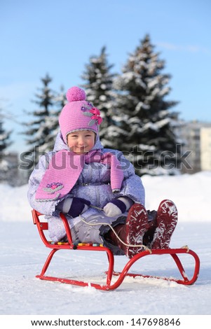Smiling little girl in pink scarf and hat sits on sled on snow in winter.