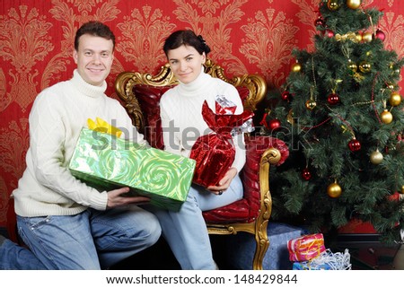 Husband and wife with gifts smile near Christmas tree in red room.