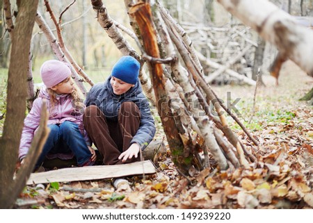 Little boy and girl sit in hut built between birches in autumn park and talk
