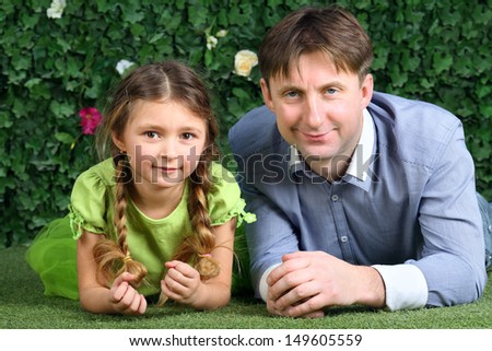 Smiling father and little daughter lie on green lawn near hedge in garden.