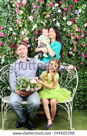 Smiling family of four sit on white bench with bunch of flowers and stand behind in garden near verdant hedge.