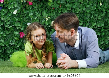 Happy father and little daughter lie on grass near hedge with flowers in garden.