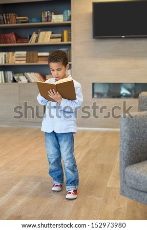 Little mulatto boy in a striped jacket and sneakers read a book in library