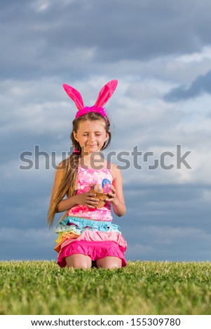 Little girl with rabbit ears on her head and with Easter eggs in hand is sitting on the grass with eyes closed