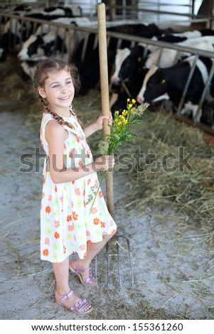 Little girl with flowers and hayfork stands at cow farm and looks away.