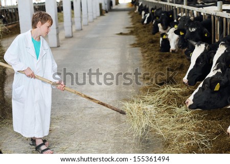 Smiling boy in white coat loads hay by big pitchfork for cows at large farm.