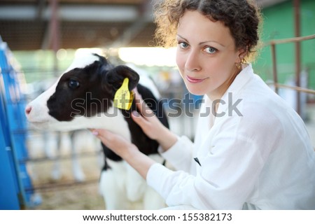 Smiling woman in white robe with small calf looks at camera in big cow farm. Focus on woman.