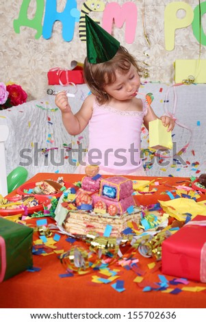 Little girl in cap drinks juice at table with cake at birthday party. Inscription Happy Birthday on wall.
