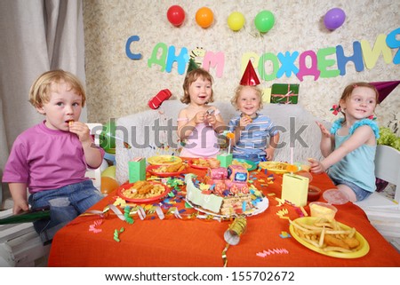 Four happy little kids eat fries with meat at red table with cake at birthday party. Inscription Happy Birthday on wall.
