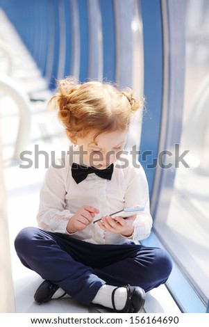Little cute girl sits near window with white cell phone in long gallery.