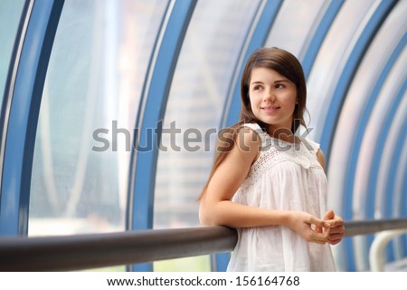 Smiling girl in white blouse stands in long blue gallery and looks in window.