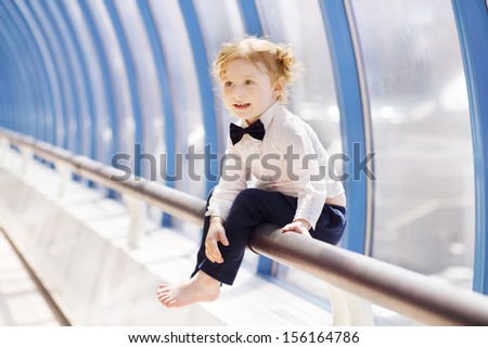 Little cute red hair girl sits on handrail in gallery near window and looks away.