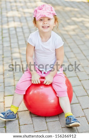 Smiling little girl sits on red ball for jumping on walkway in park
