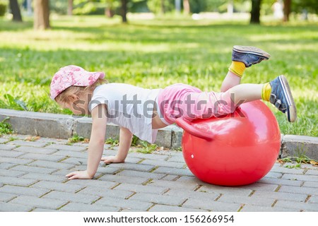 Little girl plays with red ball for jumping on  walkway in park, hands on ground, legs on ball