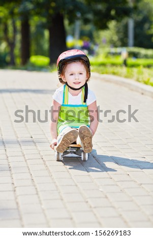 Little girl rides down park path sitting on skateboard