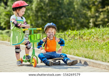 Little boy sits on skateboard with his arms akimbo ,little girl with three-wheeled scooter stands next to him
