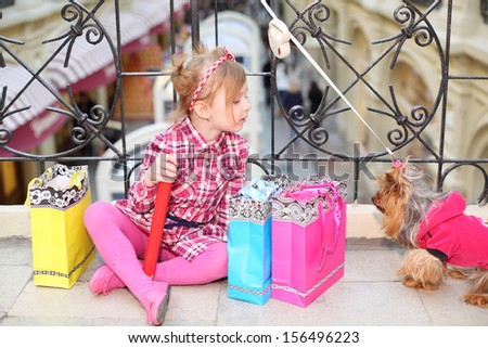 Little cute girl sits on floor near wrought railings, bags and looks at small dog in clothes in mall.