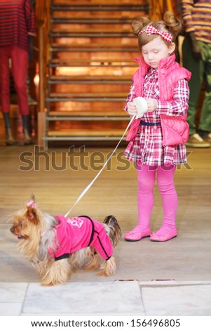 Little cute girl in pink dress leads small dog on leash in clothes in store.