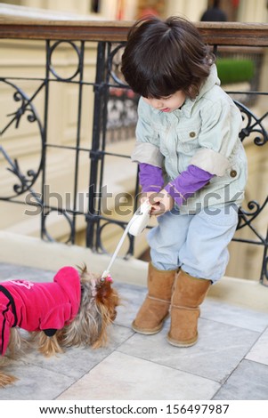 Little cute boy holds on leash small dog in pink clothes near wrought railings in mall.