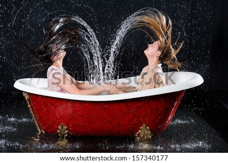 Two wet girls with closed eyes create hair splashes water in the bathtub under the spray