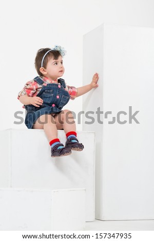 Little girl with hair band on the head is sitting on a white cube of a studio
