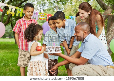 Father gives birthday cake with burning candle on it to little daughter, family of five looks at it, happy birthday sign behind their backs