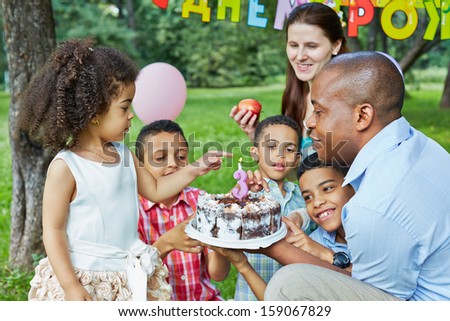 Father holds birthday cake with burning candle on it, little daughter and her three brothers finger cake, mother looks at them