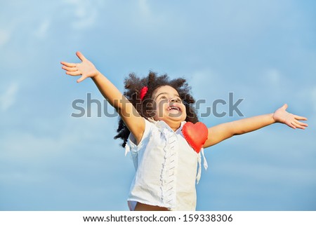 Half-length portrait of smiling little girl in white t-shirt with pinned red heart with her arms outstretched to sides and face turned to sky