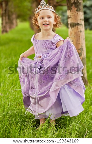 Little girl in beautiful violet gown and crown on head stands  on grassy lawn in park, lifting up slightly hem of gown