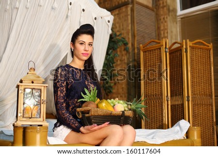 Beautiful woman sits on bed and holds basket with different fruits in bedroom.