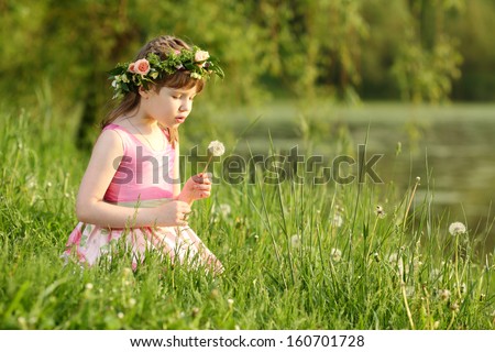 Little pretty girl in pink sits and blows on dandelion on green grass in summer park.
