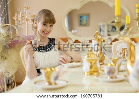 Beautiful girl in white dress sits at table with set of dishes and holds cup of tea.
