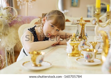 Beautiful girl in white dress sits at table with set of dishes and looks at camera.