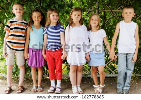 Five children stand holding hands in shadow under arch twined with green in summer park