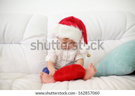 Cute little kid in red cap sits on white couch with red pillow at home. Shallow depth of field.