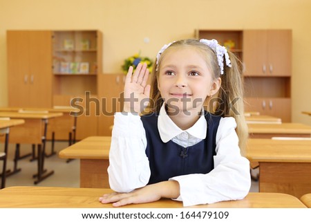 Little girl in school uniform sits at wooden school desk and raises hand to answer in classroom at school.
