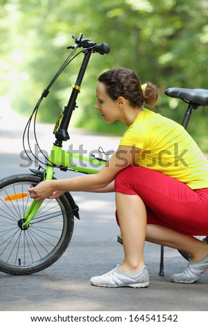 Pretty woman in sport clothes repairs bike in park at summer day.