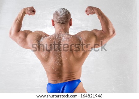 Male bodybuilder in trunks with snow on body posing back in studio