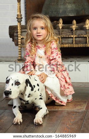 Happy pretty little girl in medieval costume sits with dalmatian near fireplace.